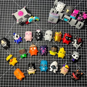 May include: A collection of small, stylized character keychains featuring various figures, including a gray treasure chest, a blue and white toy gun, and numerous colorful, cartoon-like figurines with diverse expressions and designs on a black grid.