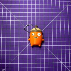 May include: A bright orange character keychain featuring a smooth, rounded body, two small arms, and wide white eyes with black pupils. The figure wears a tan, helmet-like cap with a dark brown stitched detail on top and includes a silver ball chain.