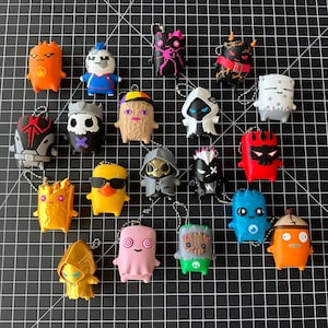 May include: A collection of twenty small, colorful, stylized character figurines, each equipped with a metal ball chain keychain attachment. The figures feature diverse designs including skulls, hooded cloaks, animals, and abstract shapes in vibrant colors.
