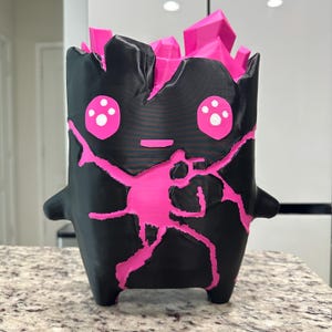 May include: A 3D-printed black creature figurine with a matte finish, featuring bright pink jagged cracks across its body, two large pink eyes with white pupils, and a cluster of sharp, geometric pink crystal-like spikes protruding from the top.