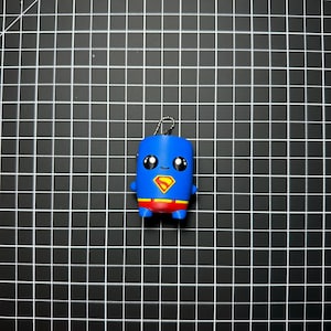 May include: A small blue figurine keychain featuring a stylized character with large black eyes, a red and yellow chest emblem, and red shorts. The figure has a smooth, rounded shape and includes a silver metal ball chain attached to the top.