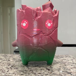 May include: A 3D printed figurine shaped like a blocky creature with a jagged, crystal-like top. The body features a gradient transition from a deep pink at the top to a vibrant green at the base, with two pink eyes containing white paw prints.