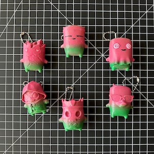 May include: Six small, pink and green gradient figurines with metal ball chains attached to the top. Each figure features a distinct character design, including one with a hood, one with a crown, one with sunglasses, and one with spiral eyes.