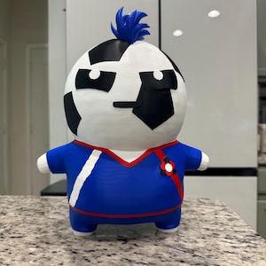 May include: A 3D printed figurine featuring a round head with black and white soccer ball patterns and a blue spiky tuft of hair. The body is a blue shirt with red trim, a white diagonal stripe, and a small red and black emblem on the chest.
