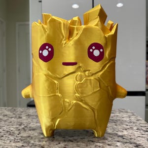 May include: A golden, 3D-printed figurine with a textured, rock-like surface and jagged top edges. The figure features a smooth, flat front with two large, dark pink eyes containing white highlights and a small, horizontal dark pink line for a mouth.