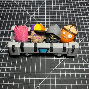 May include: A gray 3D-printed rectangular crate with black accents and a blue center detail holding four small character figurines. The figures include a pink textured creature, a tan peanut-shaped character with a colorful cap, a gray hooded skull figure, and an orange fish-like character with a brown cap.