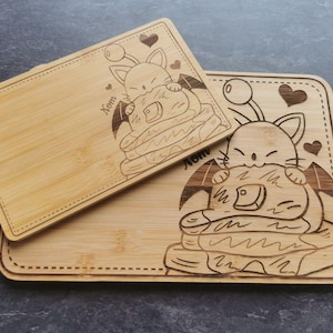May include: Two rectangular bamboo chopping boards. Each board features a laser-etched design of a cat holding pancakes. The word "Xom" is etched on each board. The boards are made of light-colored wood.