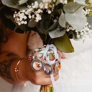 May include: A bridal bouquet featuring white roses, eucalyptus, and baby's breath. The bouquet is held with a hand adorned with a gold bracelet and a charm bracelet with photo pendants. The bouquet is tied with white and light blue ribbons.