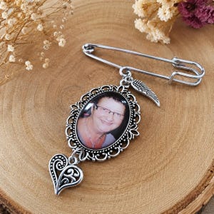 Può includere: Una spilla da balia color argento con un ciondolo ovale in una cornice filigranata decorata. Il ciondolo contiene la fotografia di una persona sorridente con gli occhiali. Un piccolo ciondolo a forma di ala e uno a cuore pendono.