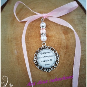 May include: A pendant with a silver-colored filigree frame, featuring the text "Gr&eacute;gory, pour toujours aupr&egrave;s de moi" on a white background. The pendant is adorned with pink and white beads and a pink ribbon.