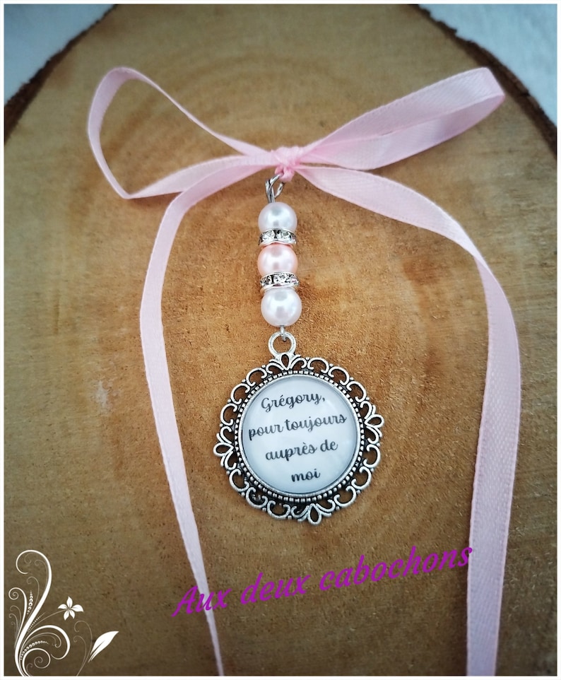 May include: A pendant with a silver-colored filigree frame, featuring the text "Gr&eacute;gory, pour toujours aupr&egrave;s de moi" on a white background. The pendant is adorned with pink and white beads and a pink ribbon.