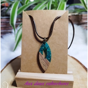Peut inclure: Un collier avec un pendentif en bois de couleur turquoise et marron, sur un cordon en cuir marron. Le pendentif a la forme d'une larme et présente un motif de grain de bois. Le texte "Aux deux cabochons" est visible sur le support en bois.