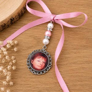May include: A round, silver-toned pendant with a photo of a person, suspended from a pink ribbon tied in a bow. The pendant is adorned with white and pink beads. The background is a wooden surface.
