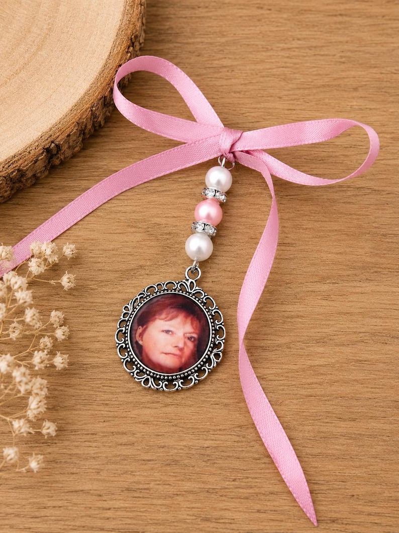 May include: A round, silver-toned pendant with a photo of a person, suspended from a pink ribbon tied in a bow. The pendant is adorned with white and pink beads. The background is a wooden surface.