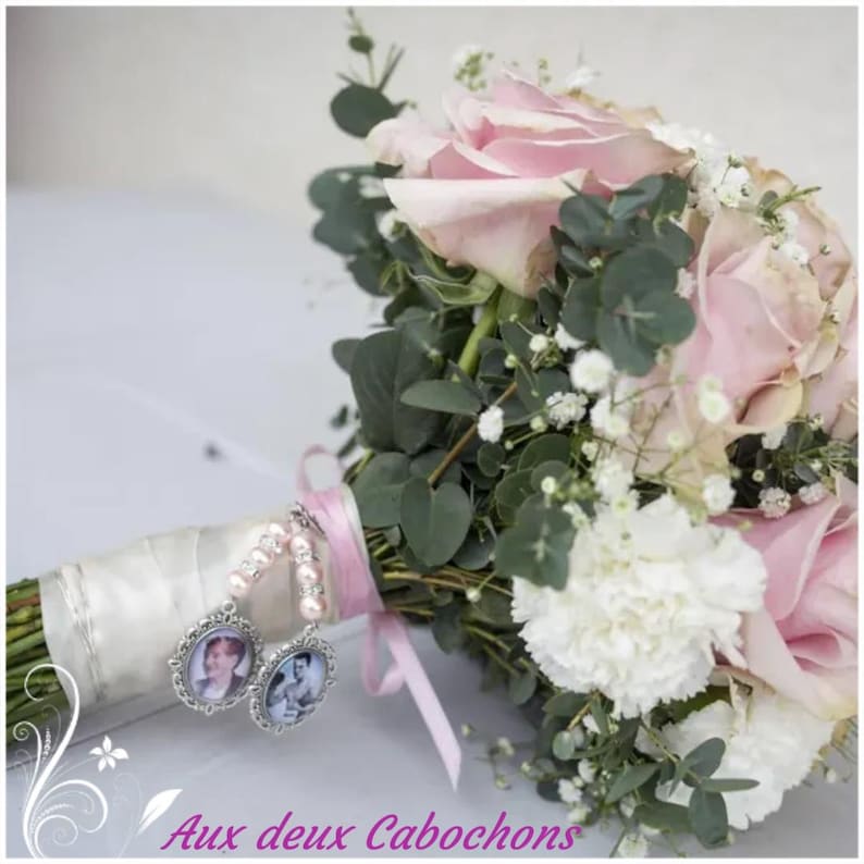 May include: A bridal bouquet featuring pink roses, white carnations, and greenery. The bouquet is adorned with two silver photo charms and tied with a satin ribbon. The text "Aux deux Cabochons" is visible at the bottom.