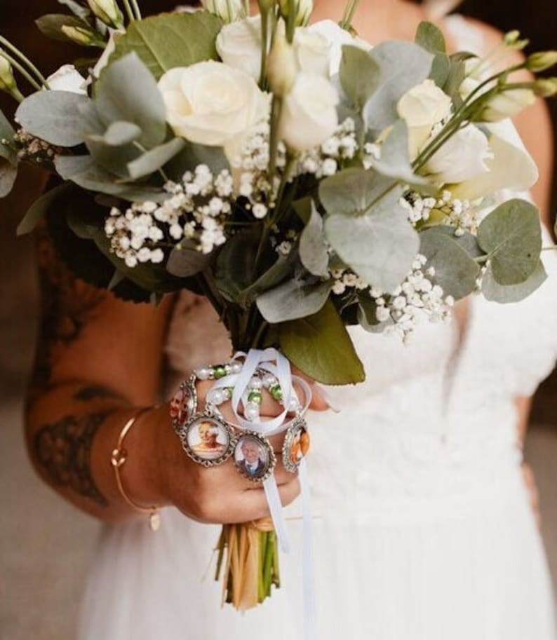 May include: A bridal bouquet featuring white roses, eucalyptus, and baby's breath. The bouquet is held with a hand adorned with a gold bracelet and a charm bracelet with photo pendants. The bouquet is tied with white and light blue ribbons.