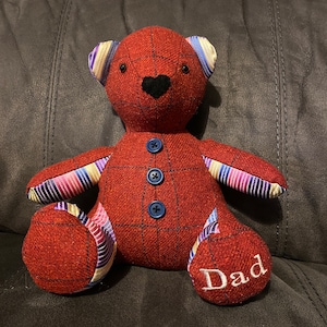 May include: A red and plaid teddy bear with blue buttons and striped fabric accents. The bear is sitting on a couch and has the word "Dad" embroidered on one foot.