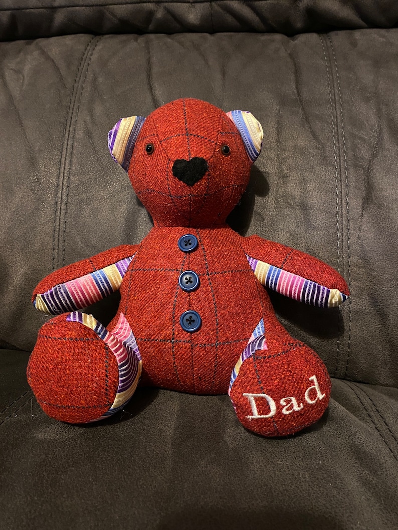 May include: A red and plaid teddy bear with blue buttons and striped fabric accents. The bear is sitting on a couch and has the word "Dad" embroidered on one foot.