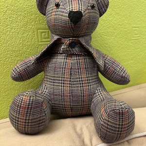 May include: A plaid teddy bear with a black nose and brown eyes. The bear is wearing a plaid shirt with a blue button.