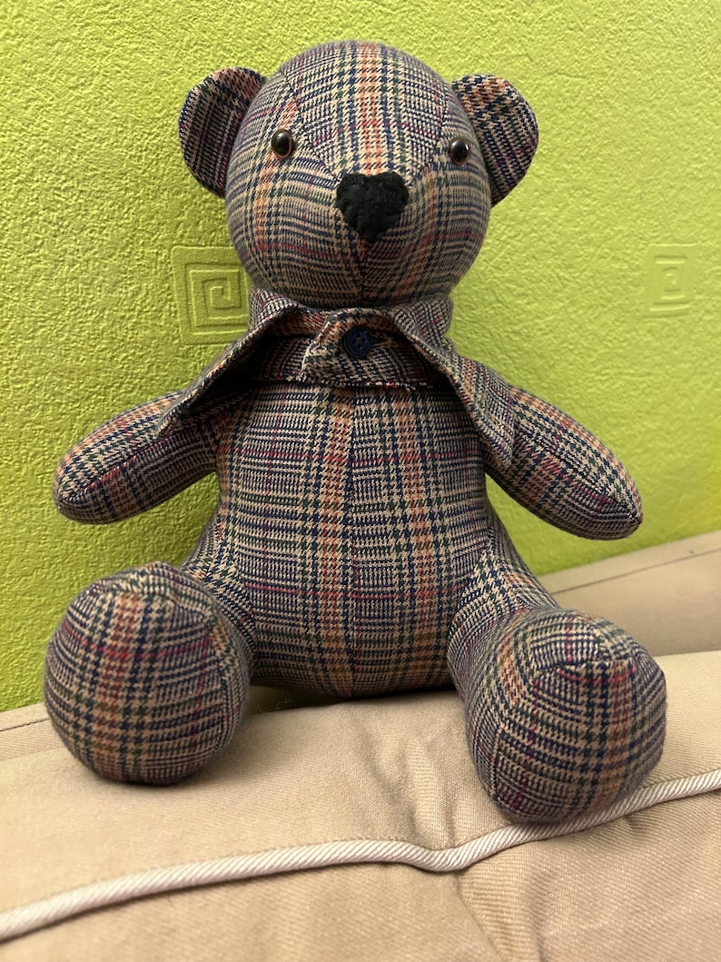 May include: A plaid teddy bear with a black nose and brown eyes. The bear is wearing a plaid shirt with a blue button.
