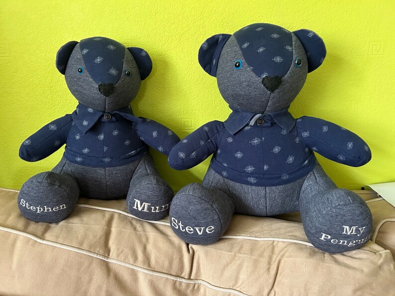 May include: Two blue teddy bears with embroidered names on their feet. The bears are wearing blue shirts with a white pattern. The bears are sitting on a light brown surface.