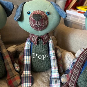 May include: A handmade stuffed dog with a teal head and denim ears. The dog has a plaid snout and collar, with the word "Pops" embroidered on its green sweater. The dog's legs are made of plaid fabric.