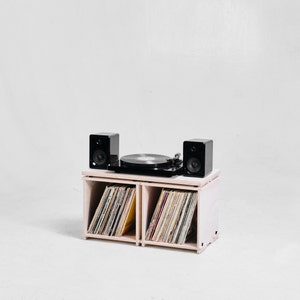 May include: A white wooden record player stand with two shelves, each holding a stack of vinyl records. The stand has a black turntable and two black speakers on top.