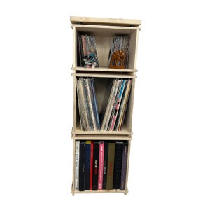 May include: A three-tiered wooden record storage unit with a light wood finish. The unit is filled with vinyl records and has a shelf for books. The top shelf has a small skull and elephant figurine.