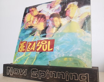 May include: A wooden record stand with the text "Now Spinning" engraved on the front. A vinyl record with a colorful image of people in water and the text "De La Soul" is displayed on the stand.