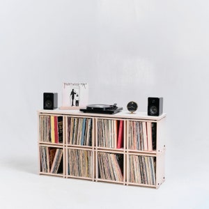 May include: A white record player shelf with six compartments, each filled with vinyl records. Two black speakers are on either side of the shelf, and a record player sits on top. A clock is on the shelf between the speakers.
