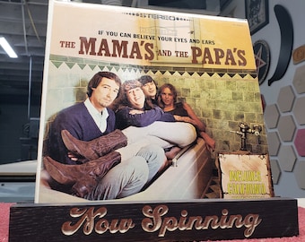 May include: A vintage vinyl record album cover for The Mamas & the Papas, displayed in a wooden stand that reads "Now Spinning." The album cover features a group of four people in a bathtub. The album is titled "If You Can Believe Your Eyes and Ears."