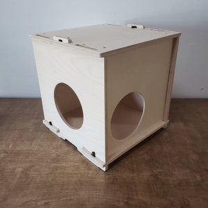 May include: A light-colored wooden cat house with two large circular openings on the sides. The box-shaped structure has a flat top with small cutouts and is set on a wooden surface.