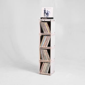 May include: A white vinyl record storage unit with four shelves. The shelves are filled with vinyl records. The top shelf has a black vinyl record with the text "Elton John - Goodbye Yellow Brick Road" on the cover.