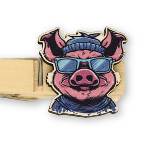 Könnte beinhalten: Eine Holzklammer mit einem rosa Schwein, das eine blaue Sonnenbrille und eine blaue Mütze trägt. Das Schwein lächelt und trägt einen blau-weiß gestreiften Schal.