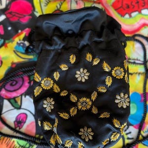 May include: Black satin drawstring bag with gold floral embroidery. The bag features a black rope strap and is set against a colorful, patterned background. The floral design includes gold leaves and silver-toned accents.