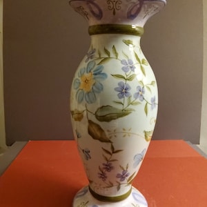 May include: A white ceramic vase with a light green and blue floral design. The vase has a scalloped rim and a wide base.
