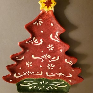 May include: A red ceramic Christmas tree-shaped plate with white floral and dot details. The tree is topped with a yellow star with a red centre and has a green base. A decorative piece for festive dining during the holiday season.