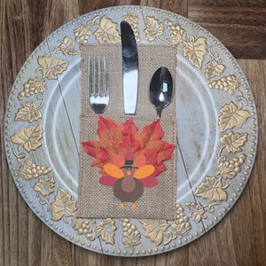 May include: A round decorative plate with gold leaf and grape motifs features a burlap utensil holder. The holder contains a silver fork, knife, and spoon, and is decorated with artificial autumn leaves and a cartoon turkey wearing a hat.