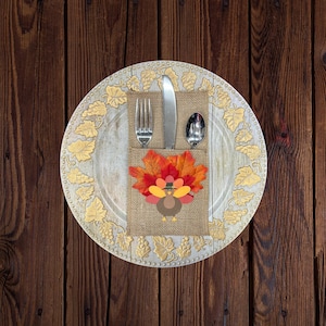 May include: Thanksgiving table setting with a decorative plate, silverware, and a burlap napkin. The napkin features a turkey design with fall leaves. The plate has a gold leaf design around the edge, set on a wooden table.
