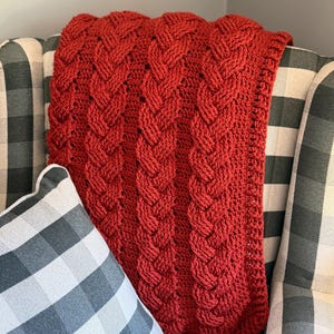 May include: A deep red crochet blanket featuring a prominent vertical cable knit pattern. The textile displays a thick, textured weave with repeating braided columns that run the length of the fabric, creating a raised, dimensional surface design.