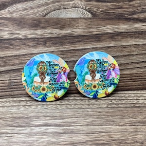 May include: Two round car coasters with a floral design. The coasters feature a woman with a sunflower crown and the text "They whispered to her, you can't withstand the storm. She whispered back, I am the storm."