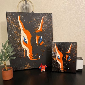 Puede incluir: Dos pinturas de lienzo negro con diseños abstractos naranja y azul de la cara de un zorro. Las pinturas están sobre una superficie negra con una pequeña figura roja y naranja delante de ellas.