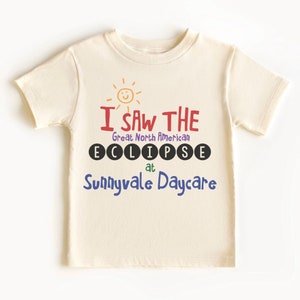 May include: A cream-coloured t-shirt featuring the text "I SAW THE Great North American ECLIPSE at Sunnyvale Daycare." The text is in various colours, including red, purple, and blue, with a smiling sun graphic above.