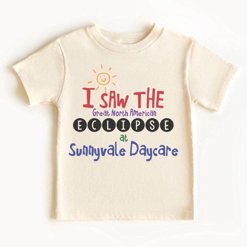 May include: A cream-coloured t-shirt featuring the text "I SAW THE Great North American ECLIPSE at Sunnyvale Daycare." The text is in various colours, including red, purple, and blue, with a smiling sun graphic above.