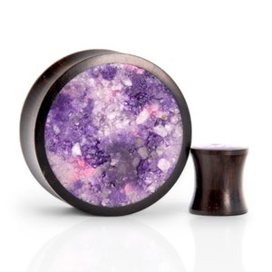 May include: A pair of wooden ear gauges with a purple and pink speckled design. The larger gauge is round and the smaller gauge is cylindrical.