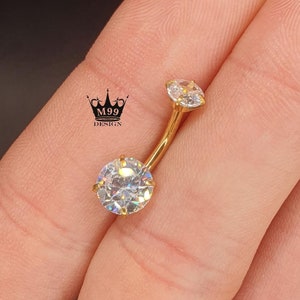 Pode incluir: Anel de umbigo dourado com duas pedras redondas de zircônia cúbica transparentes.