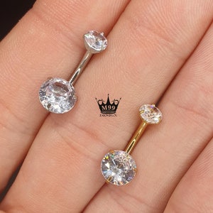 Puede incluir: Dos anillos de ombligo con piedras brillantes transparentes. El anillo de la izquierda es plateado y el anillo de la derecha es dorado.