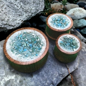 May include: Three wooden ear gauges with a blue and white crystal design. The gauges are different sizes and are arranged in a row.