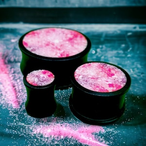 May include: Three black wooden ear gauges with pink and white speckled designs. The gauges are arranged in a pyramid shape on a blue surface.