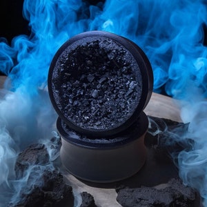 Pode incluir: Um par de expansores de orelha de madeira preta com um design texturizado preto e cinza. Os expansores estão sobre uma superfície de madeira com fumaça azul no fundo.